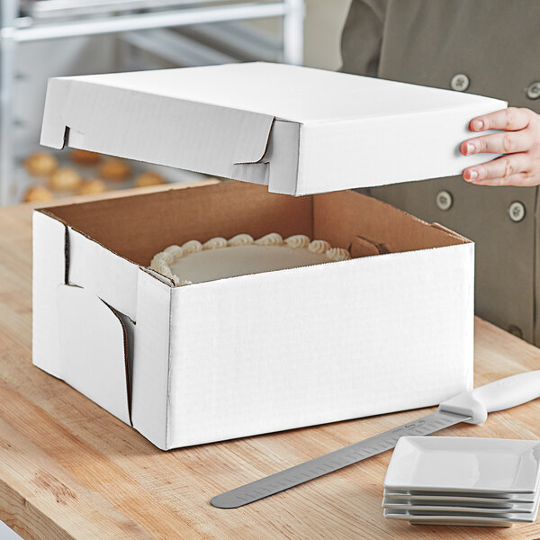 A hand opening a white bakery box to reveal a white cake.