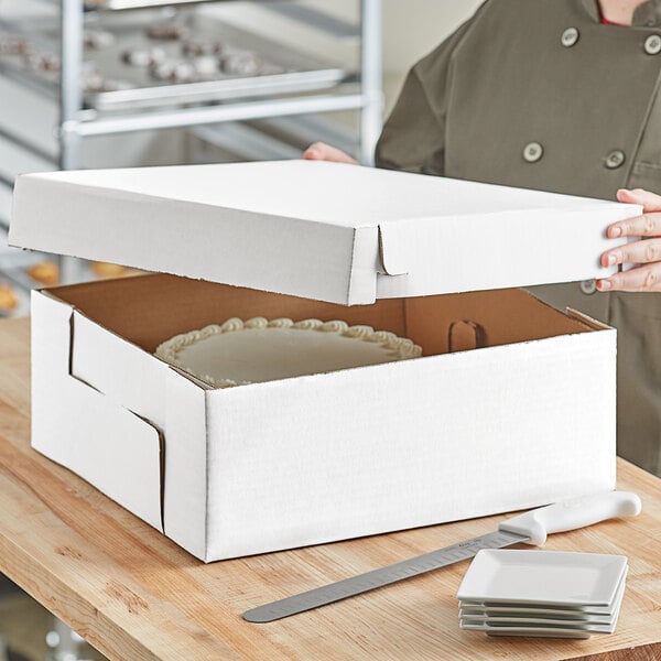 A woman opening a white corrugated bakery box with a white cake inside.