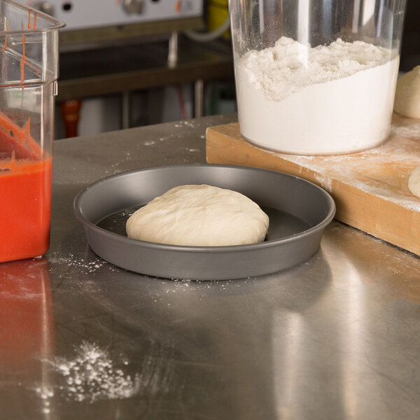 A dough in an American Metalcraft hard coat anodized aluminum pizza pan on a counter.