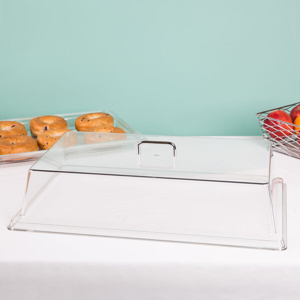 A clear Cambro display cover over a tray of donuts with a basket of fruit on a counter.