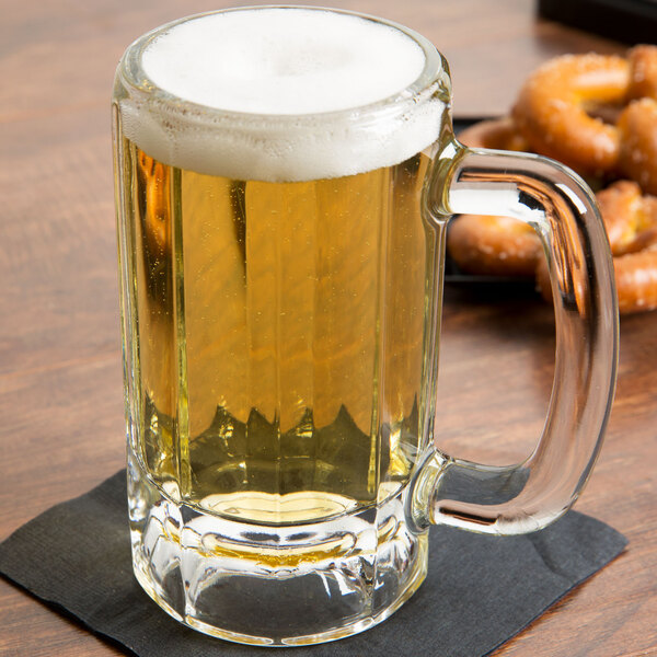A Libbey paneled beer mug filled with beer with foam on top on a napkin with pretzels.