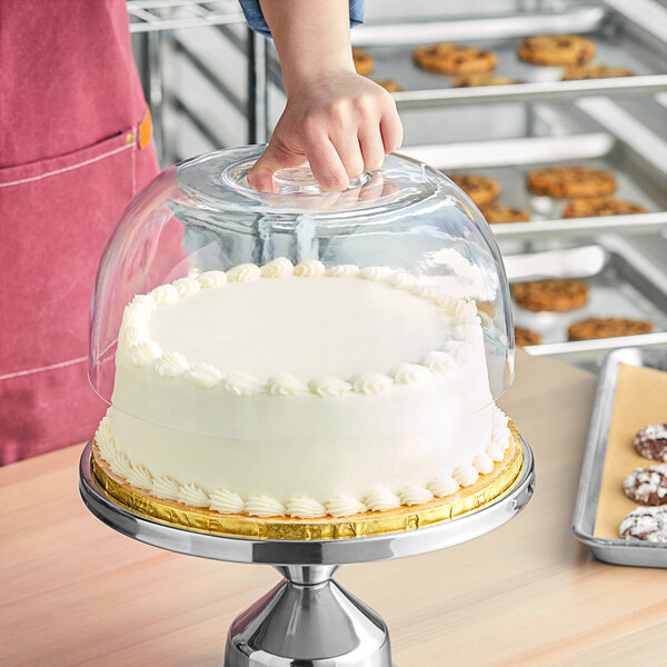A person putting a white cake on a stand with a Tablecraft clear dome cover.