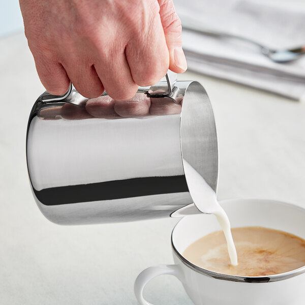 A person pouring milk into a cup of coffee using a Tablecraft stainless steel frothing pitcher.