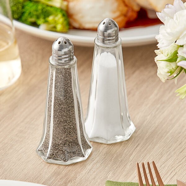 An American Metalcraft glass salt shaker on a table with a plate of food.