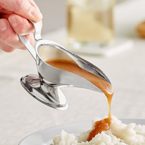 A person pouring gravy into a bowl of mashed potatoes from an American Metalcraft stainless steel gravy boat.
