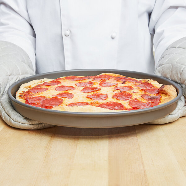 A person holding a pepperoni pizza in an American Metalcraft hard coat anodized aluminum pizza pan.