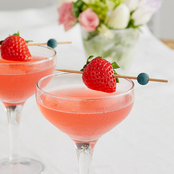 Two glasses of pink liquid with a strawberry and a Front of the House Green Bamboo Ball Pick on the rim of each glass.