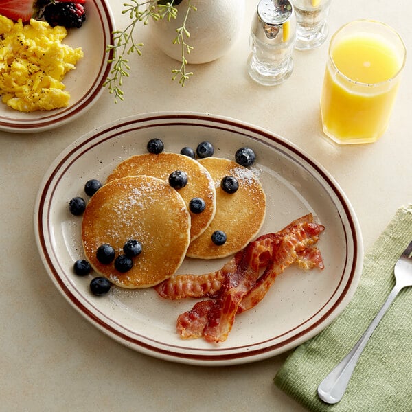 A brown speckled narrow rim oval stoneware platter with a stack of pancakes topped with blueberries and bacon.