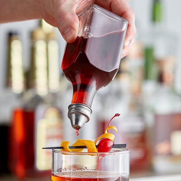 A hand using a Choice clear glass bitters bottle with a stainless steel pourer to pour a red liquid into a glass.
