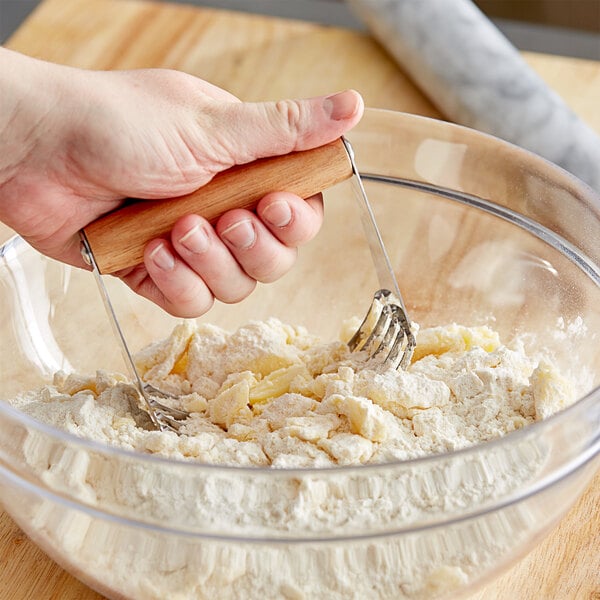 A hand using a Choice stainless steel pastry blender with a wood handle to mix dough in a bowl.