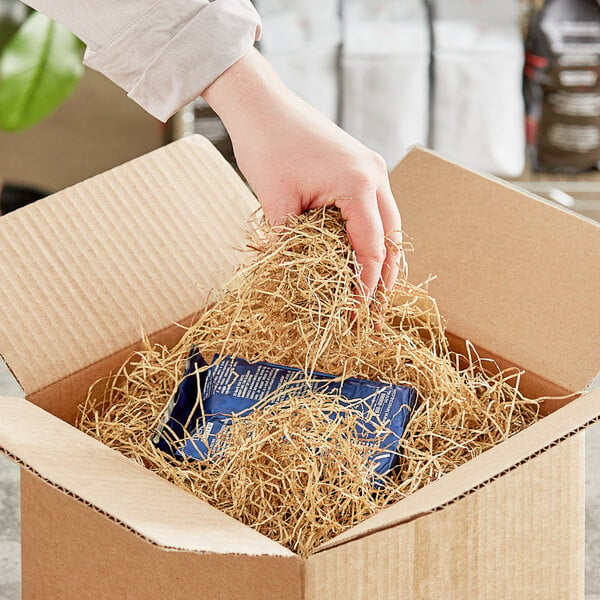 A hand holding a blue package in a cardboard box filled with Spring-Fill Kraft Very Fine Paper Shred.