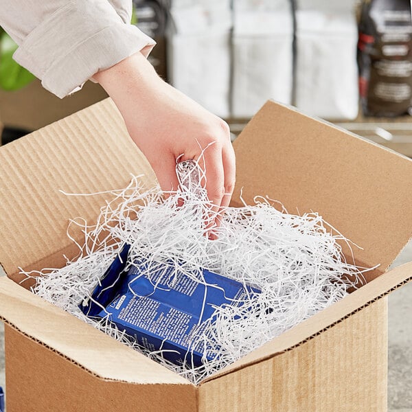 A hand putting a blue box of Spring-Fill Very Fine Paper Shred into a cardboard box filled with shredded paper.
