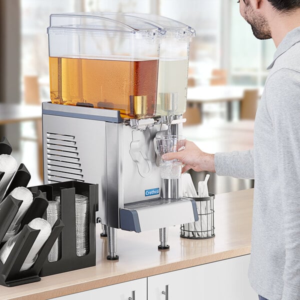 A man pouring liquid into a Crathco mini double beverage dispenser on a counter in a professional kitchen.