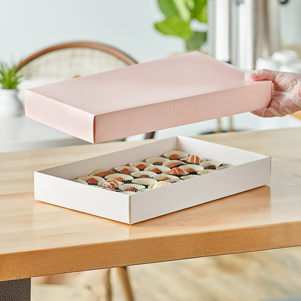 A hand holding a pink 14 3/4" x 9" x 2" candy box on a table with chocolates inside.