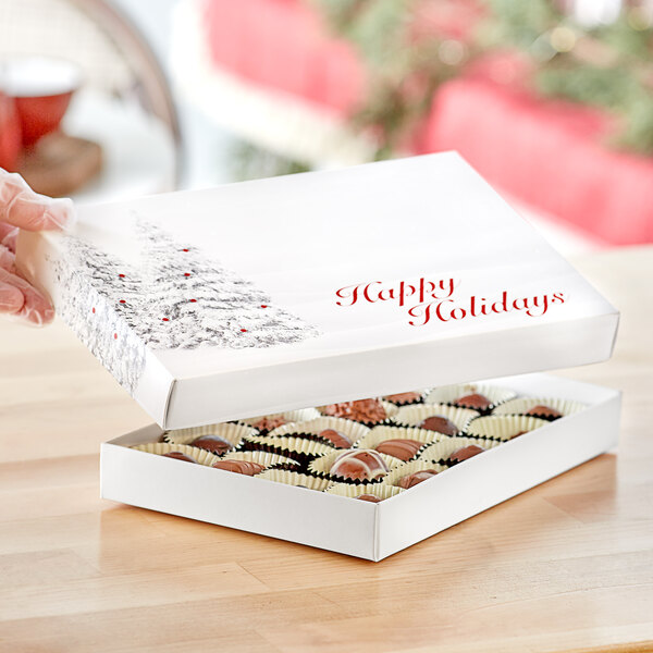 A hand holding a white Happy Holidays candy box with red text and a tree on it containing chocolates.