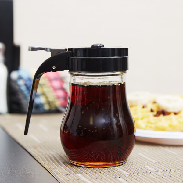 A Tablecraft glass syrup dispenser with a black top filled with red syrup on a table.