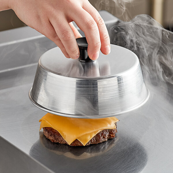 A hand holding a American Metalcraft aluminum basting cover over a cheeseburger.