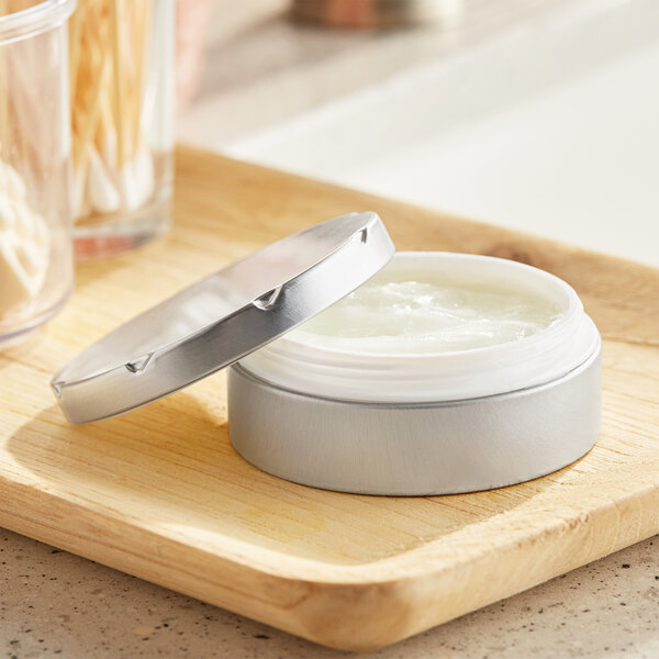 A silver metal tin with a notched lid and plastic ring holding a white jar of cream.