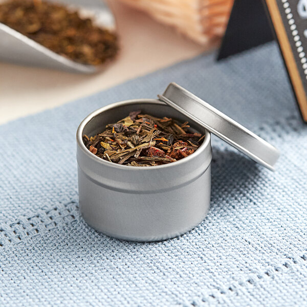 A silver metal tin with a slip cover full of brown herbs.