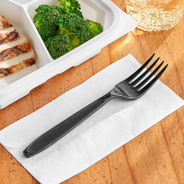 A Solo Impress black plastic fork on a napkin next to a plate of broccoli.