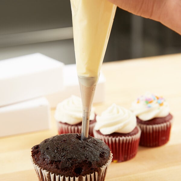 A person using an Ateco small bismark piping tip in a pastry bag to frost a chocolate cupcake with white icing.