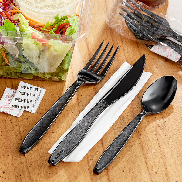 A wrapped Solo Impress black plastic fork and knife with a napkin on a wooden surface next to a salad.