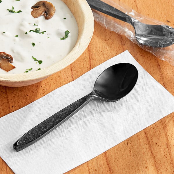 A Solo Impress heavy weight black plastic soup spoon wrapped in plastic on a table with a bowl of soup and a napkin.