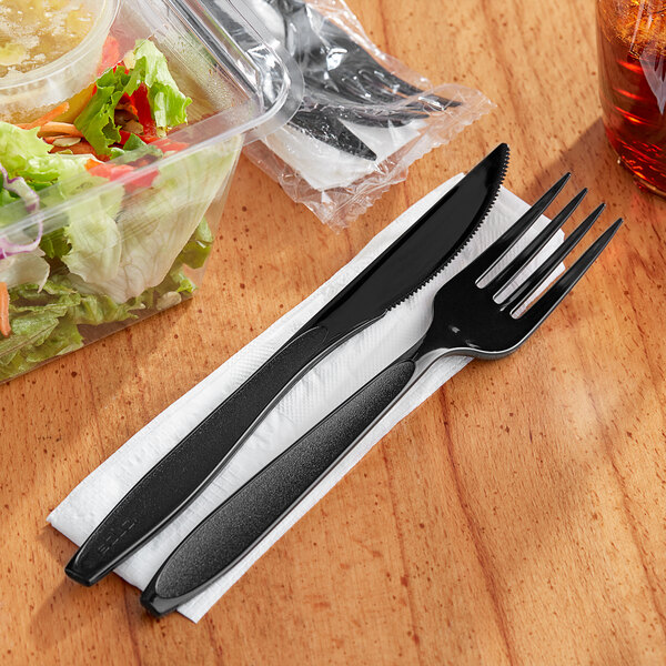 A Solo Impress black plastic fork and knife on a napkin next to a salad in a plastic container.