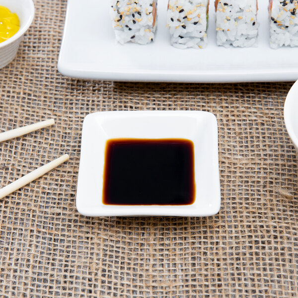 A square white porcelain sauce cup with black liquid on a plate of sushi.