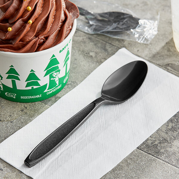 A black Solo Impress plastic teaspoon on a white napkin with a cup of chocolate ice cream.