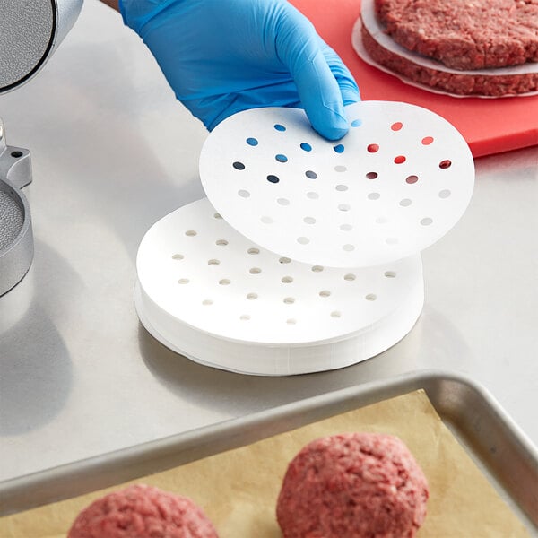 A person in a blue shirt and gloves preparing hamburgers on a counter using white round perforated paper.