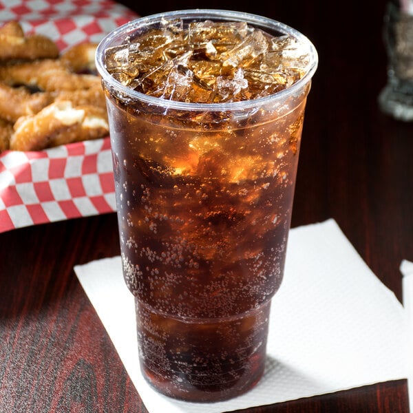 A Solo Ultra Clear plastic cup of soda with ice on a table with a basket of pretzels.