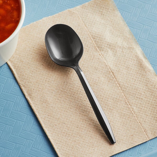 A black plastic soup spoon on a beige napkin next to a bowl of soup.