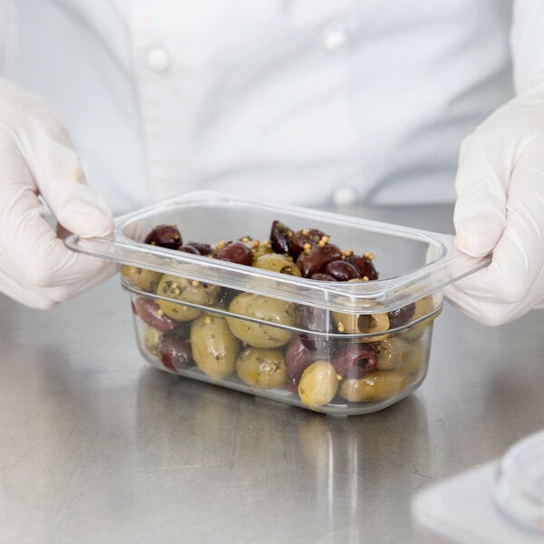 A person in gloves holding a Carlisle clear plastic food pan filled with olives.