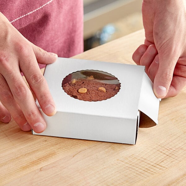 A person's hand holding a Baker's Mark white bakery box with a cookie inside.