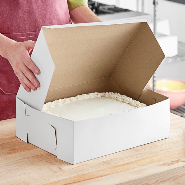 A person opening a white Baker's Mark bakery box to reveal a cake.