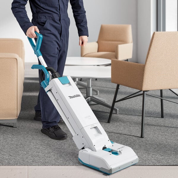 A person in overalls using a Makita cordless upright vacuum cleaner in a professional kitchen.