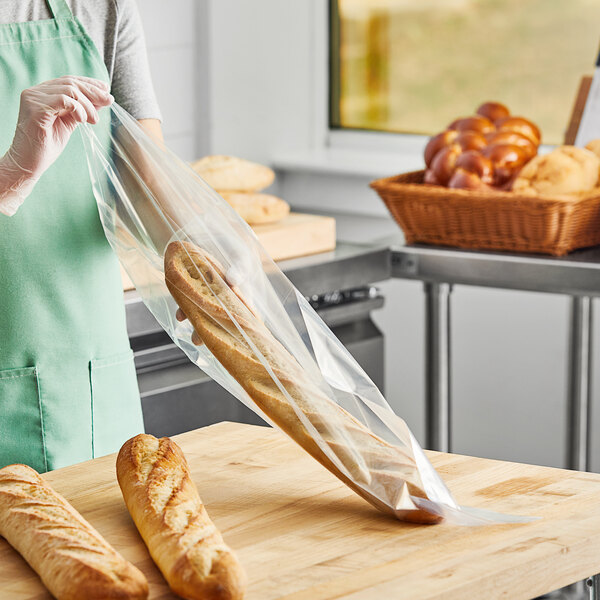 A woman in a green apron holding a Choice plastic bag with a baguette inside.
