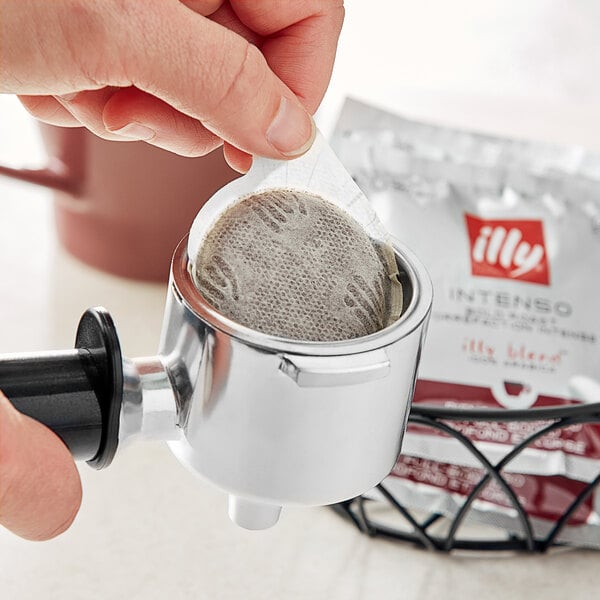 A hand using an illy Intenso espresso pod in a coffee machine.