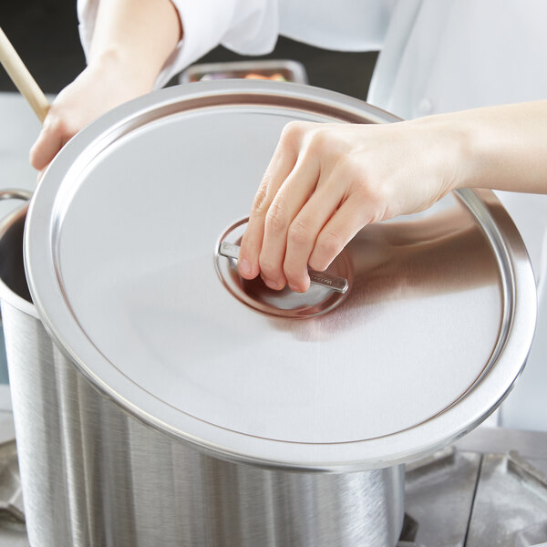A person's hand holding a silver lid over a large silver pot.