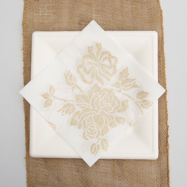 A white Hoffmaster linen-like dinner napkin with a gold floral design on a white plate.