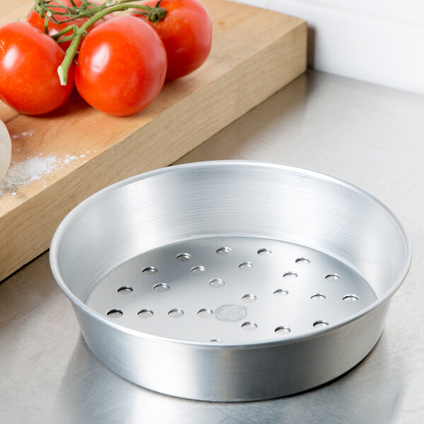 An American Metalcraft aluminum pizza pan with holes in it sitting on a white surface with a tomato close by.