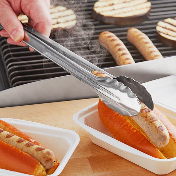 A person using Choice stainless steel utility tongs to hold a hot dog over a grill.