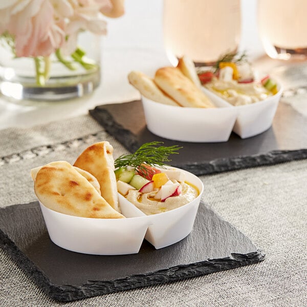 A white plastic mini dual compartment dish with hummus and vegetables on a table.
