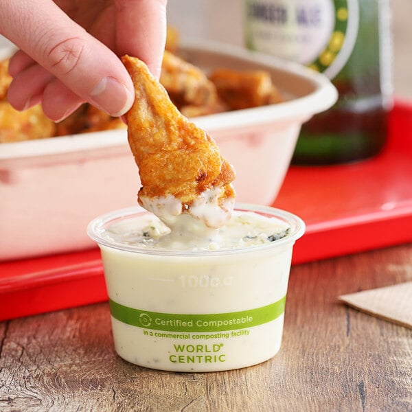 A hand holding a piece of fried chicken dipping into a World Centric compostable portion cup filled with white sauce.