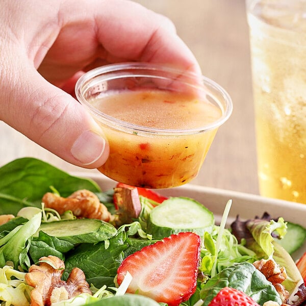 A hand holding a World Centric compostable portion cup of yellow dressing over a salad.