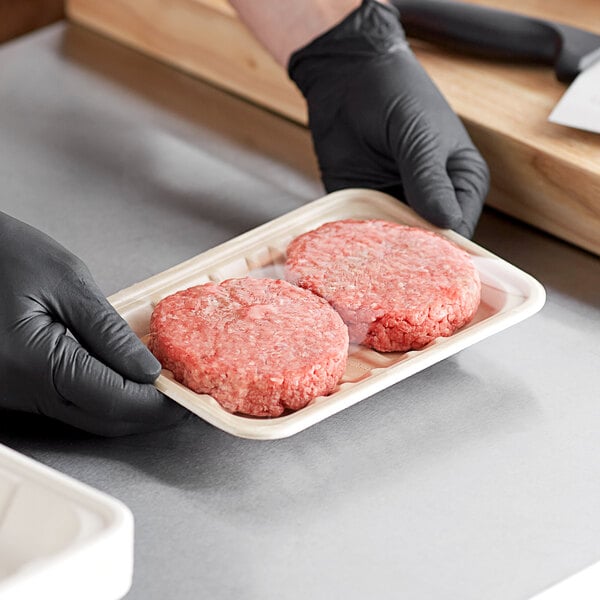 A person in black gloves holding a World Centric compostable fiber laminated meat tray with two raw hamburger patties.