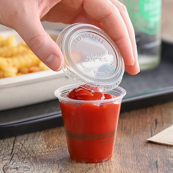 A hand holding a World Centric compostable clear plastic lid over a small plastic cup of ketchup.