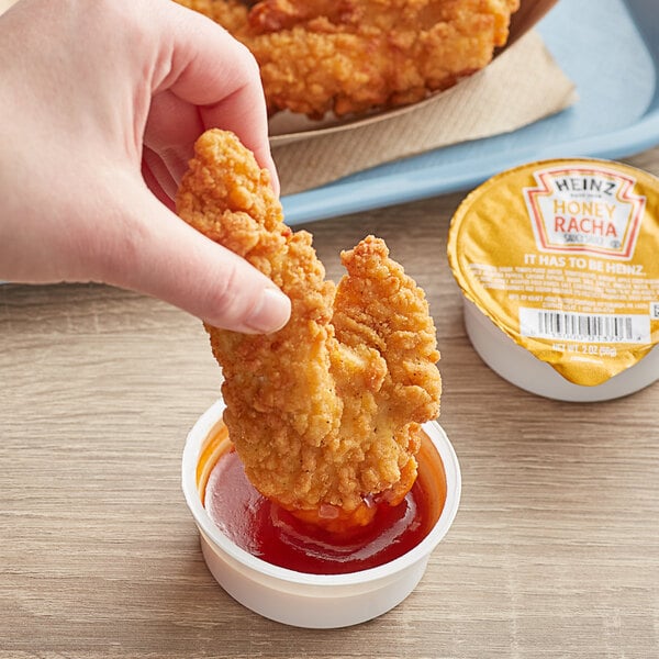 A hand holding a piece of fried chicken over a small container of Heinz Honeyracha sauce.