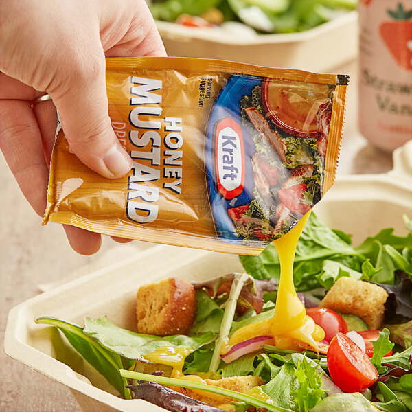 A hand pouring Kraft Honey Mustard dressing from a packet onto a salad at a salad bar.